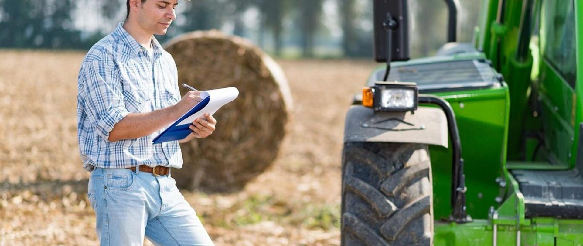 Rolnik stojący na tle pola z belą słomy, trzymający z dłoni notes do którego zapisuje informacje. Obok rolnika stoi traktor. 