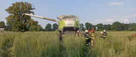 Przedstawia kombajn marki CLAAS stojący na polu w którym doszło do pożaru. Przy maszynie strażacy uczestniczący w jej gaszeniu