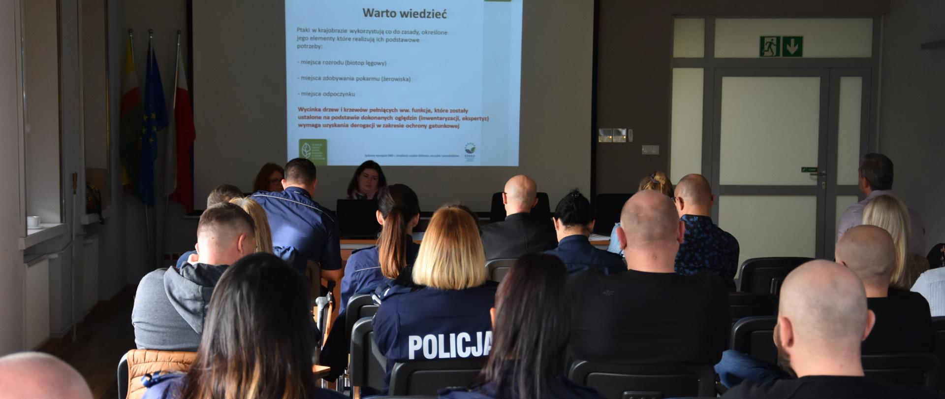 Grupa kilkudziesięciu osób siedzących tyłem. Przed nimi za stołem siedzą dwie kobiety. Za ich plecami na ścianie wisi ekran z wyświetlonym slajdem z prezentacji.