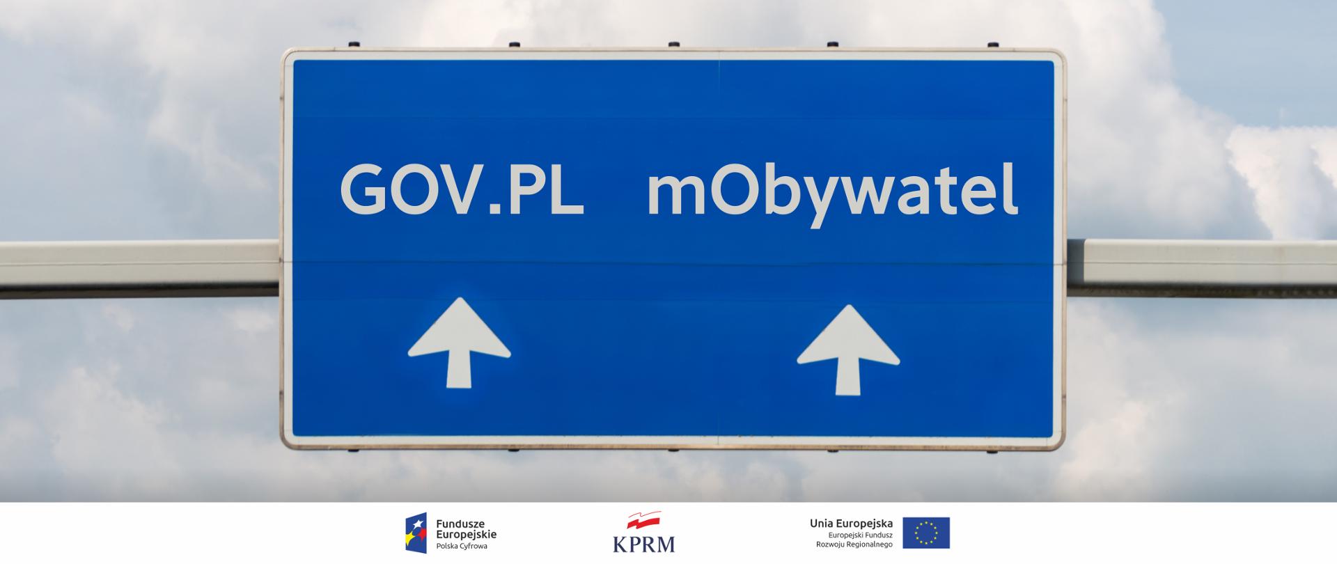 Znak drogowy (poziomy, na niebieskim tle, wskazujący kierunki, wiszący nad drogą). Na dole znaku dwie strzałki wskazujące trasę, nad lewą - napis GOV.PL, nad prawą - mObywatel.