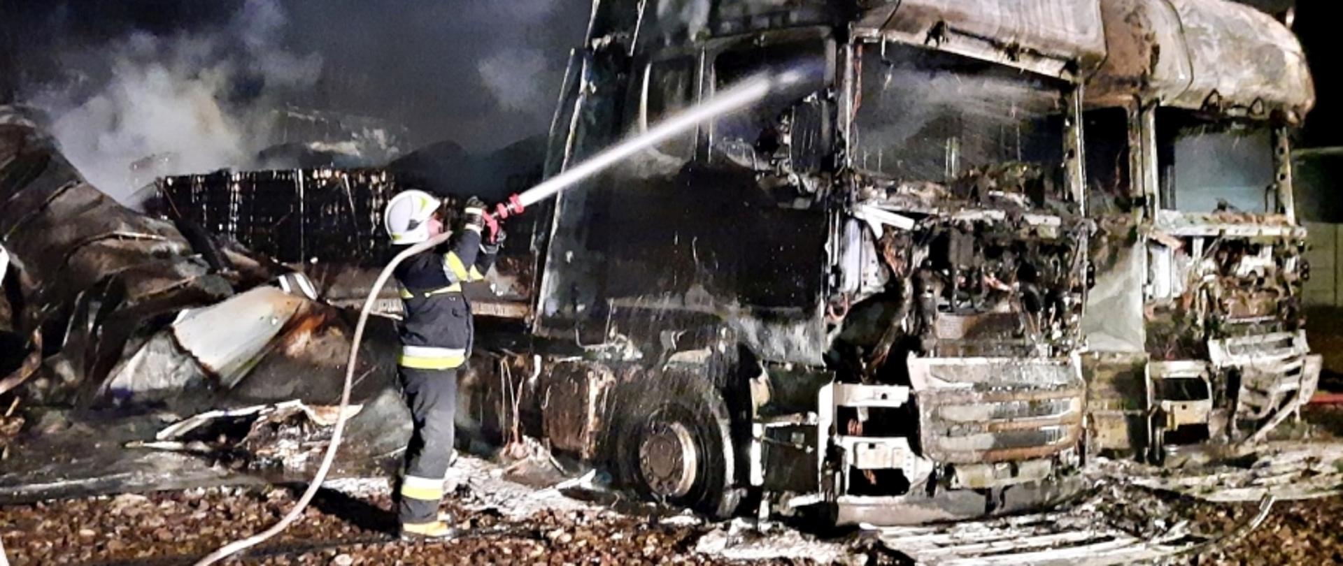 Na zdjęciu widoczny strażak OSP w białym hełmie oraz wraki dwóch ciągników siodłowych. Strażak dogasza jeden z nich prądem wody.