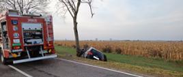 Na zdjęciu widzimy tył samochodu osobowego koloru czarnego który wjechał do rowu i uderzył w drzewo. Z lewej strony kadru widzimy samochód strażacki koloru czerwonego. Samochód strażacki stoi na asfaltowej drodze, srebrne żaluzje uniesione do góry. 