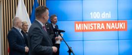 Minister Wieczorek i wiceministrowie stoją z boku sali, pod wielkim wbudowanym w ścianę ekranem z napisem 100 dni ministra nauki.