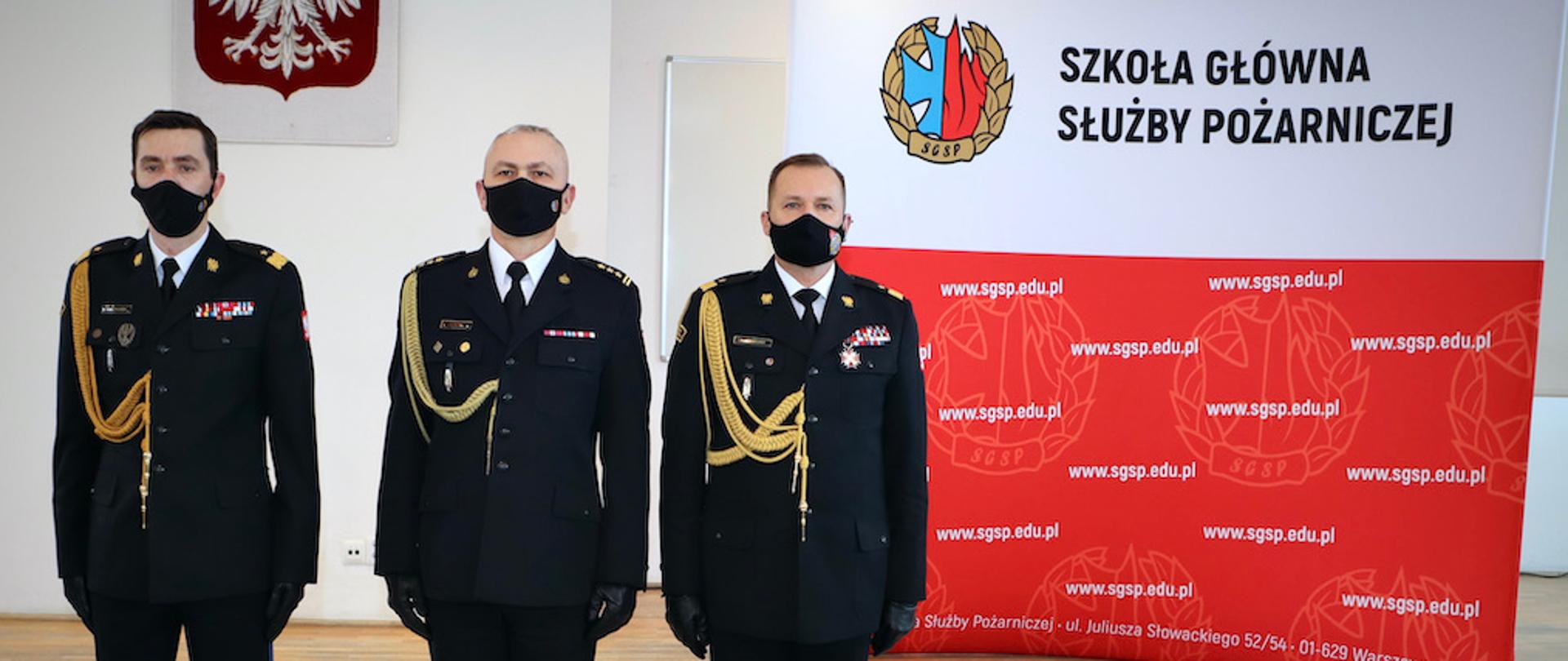 Zdjęcie pamiątkowe . Trzej mężczyźni w mundurach. W tle godło oraz baner reklamowy z napisem Szkoła Główna Służby Pożarniczej
