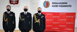 Zdjęcie pamiątkowe . Trzej mężczyźni w mundurach. W tle godło oraz baner reklamowy z napisem Szkoła Główna Służby Pożarniczej