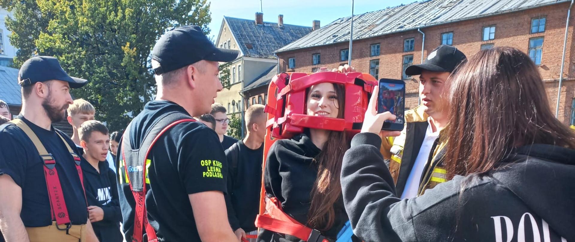 Usztywniona na desce uczennica stoi na placu manewrowym asekurowana przez strażaków