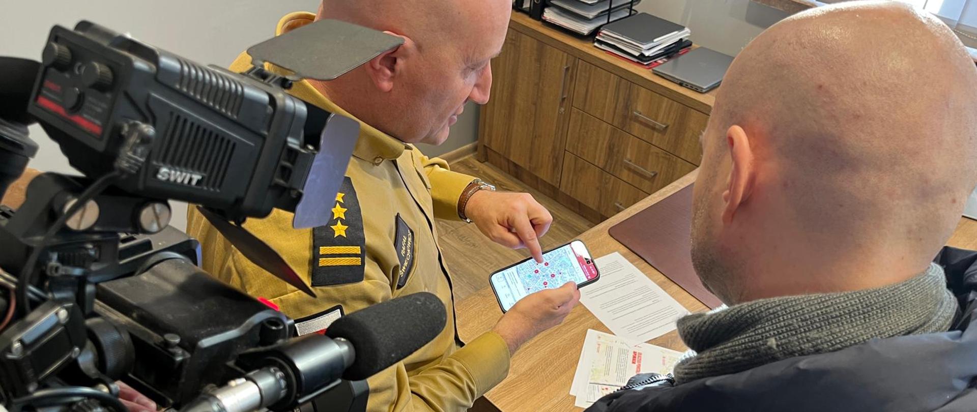 Strażak prezentuje w dłoniach telefon z aplikacją. Z prawej strony mężczyzna, przygląda się rozmówcy, z lewej zaś widać kamerę z mikrofonem.