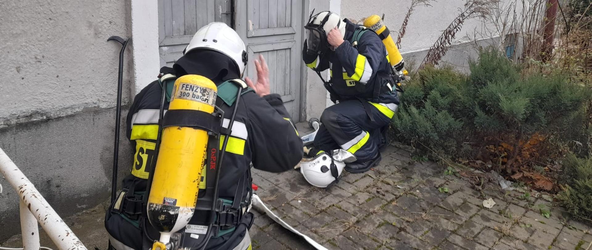 Zdjęcie z egzaminu praktycznego przedstawia dwóch strażaków ubranych w nomeksy, którzy kucają przed drzwiami, ubierają maskę do aparatu powietrznego i hełm aby zaraz wejść przez drzwi do mieszkania, w którym zasymulowano pożar.