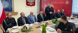 Przy stołach na których znajdują się dekoracje świąteczne i talerze, siedzą strażacy i zaproszenie goście. Strażacy ubrani są w mundury galowe zaproszeni goście odświętnie w garniturach. Zastępca Komendanta w postawie stojącej przemawia. W tle logo Państwowej Straży Pożarnej oraz godło Polski i flagi