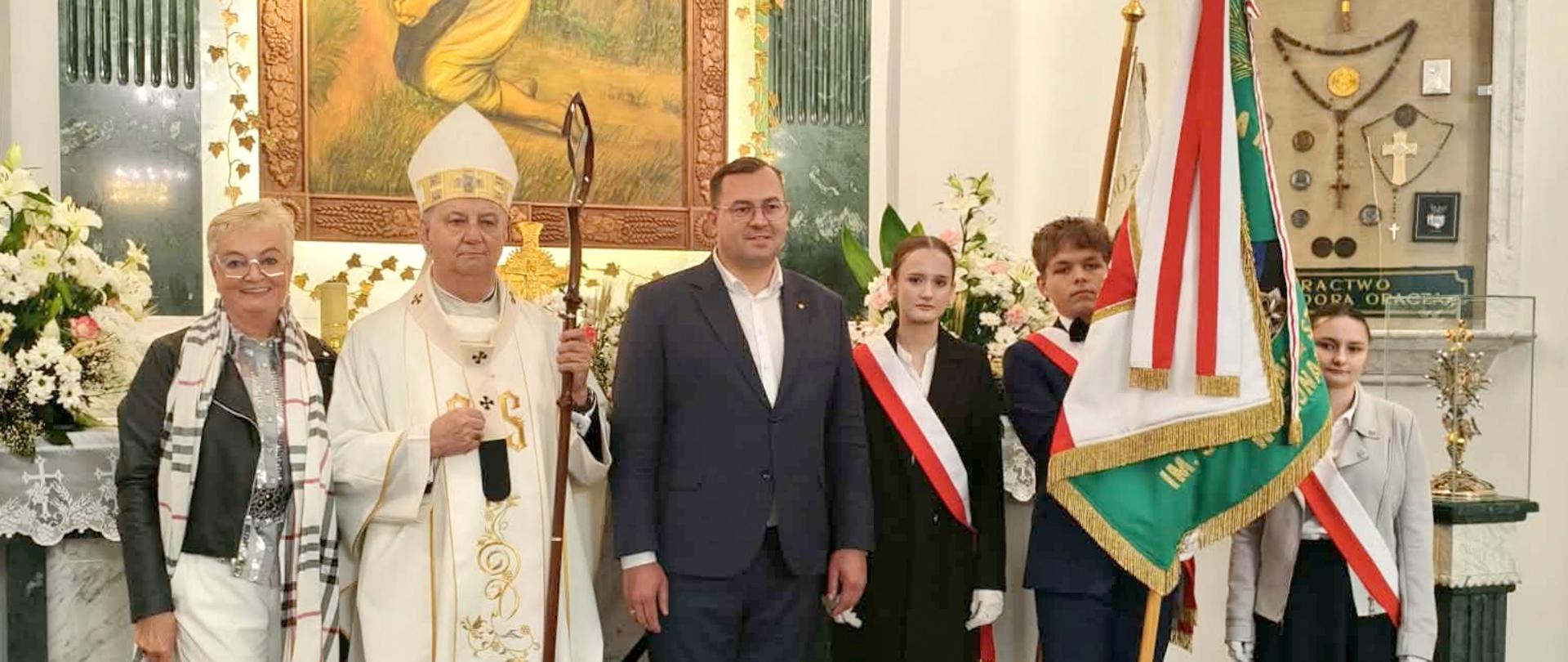Grupa osób, m.in. biskup i członkowie pocztu sztandarowego, pozują do zdjęcia na tle obrazu przedstawiającego św. Izydora Oracza.