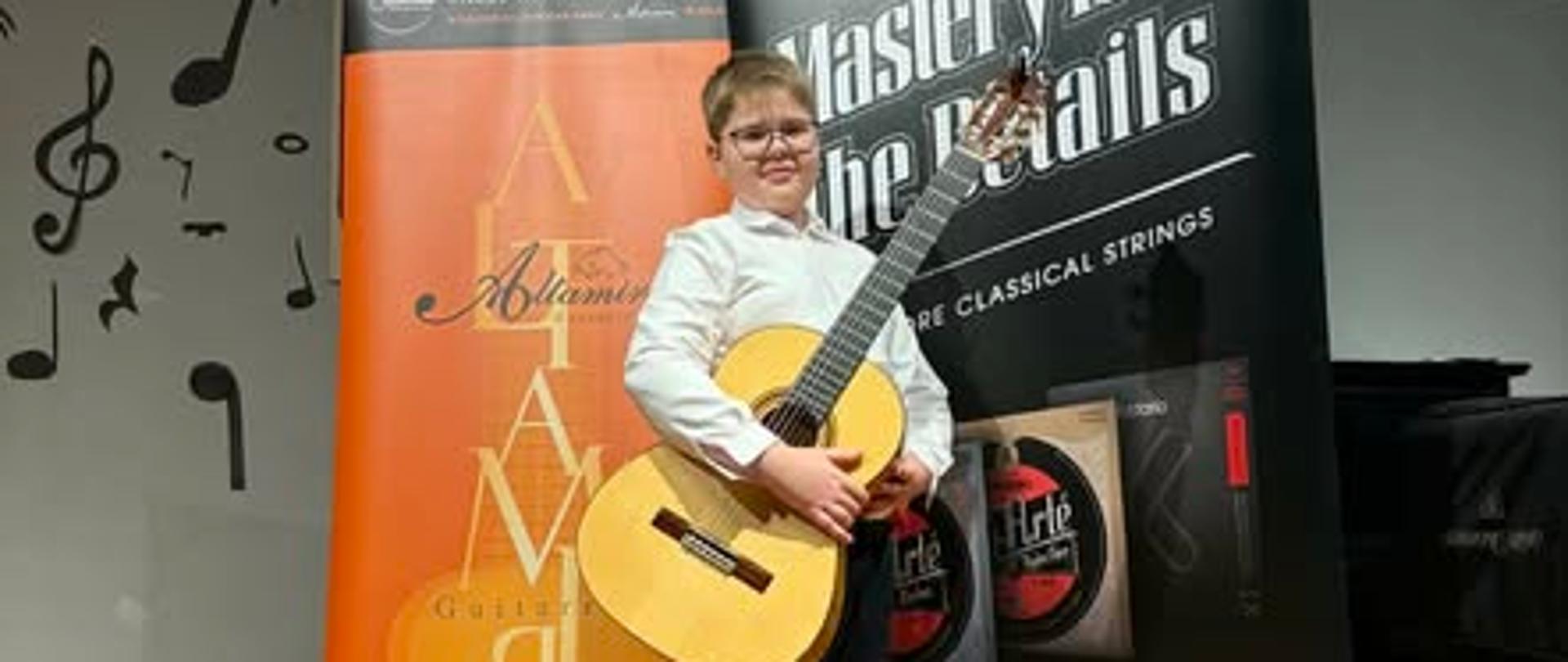 Na drewnianej podłodze stoi chłopiec ubrany w białą koszulę i czarne spodnie. W ręku trzyma gitarę. Za nim dwa rozstawione roll-upy. 