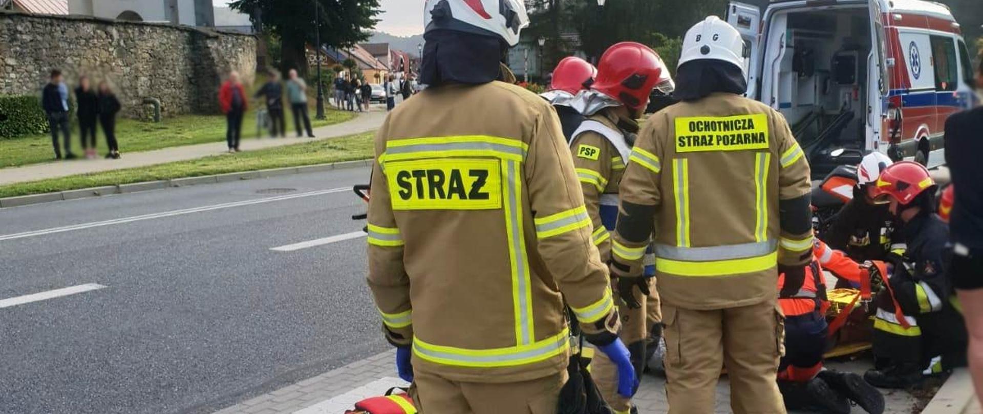 Strażacy i ratownicy medyczni udzielają pomocy osobie poszkodowanej na zatoczce autobusowej. W tle karetka Pogotowia ratunkowego.
