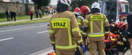 Strażacy i ratownicy medyczni udzielają pomocy osobie poszkodowanej na zatoczce autobusowej. W tle karetka Pogotowia ratunkowego.