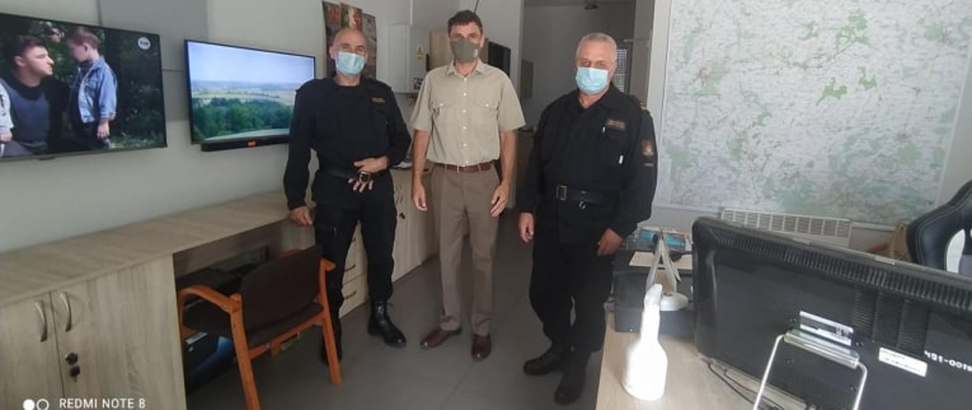 Punkt alarmowo-dyspozycyjny w Nadleśnictwie Piaski. Zastępca komendanta powiatowego PSP w Rawiczu i dowódca jednostki ratowniczo-gaśniczej w towarzystwie nadleśniczego pana Tomasz Kwiecińskiego. Z lewej strony na ścianie wisi zegar oraz monitory, z prawej mapa.