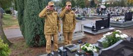 Zdjecie przedstawia dwóch strażaków w mundurach służbowych koloru musztardowego salutujących nad grobem