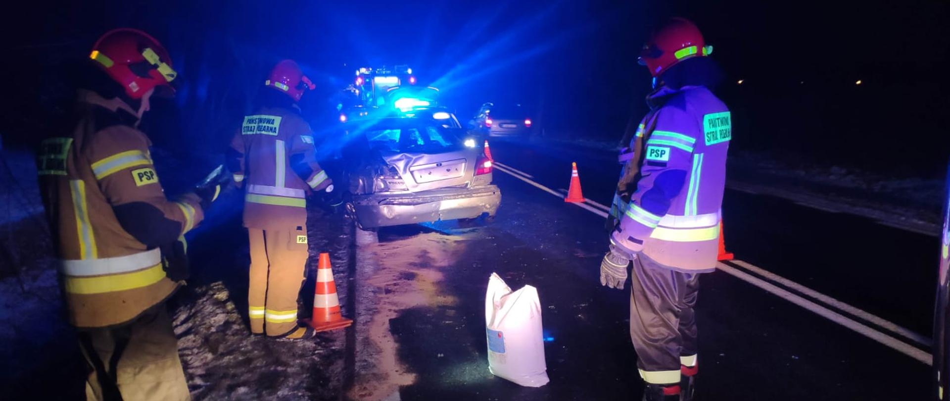 W nocy, na drodze asfaltowej stoi 3 strażaków w strojach strażackich i czerwonych hełmach, przed nimi stoi srebrny samochód osobowy ze zniszczonym tyłem w wyniku wypadku samochodowego. Przed samochodem stoi samochód strażacki i policyjny z włączonymi światłami. 