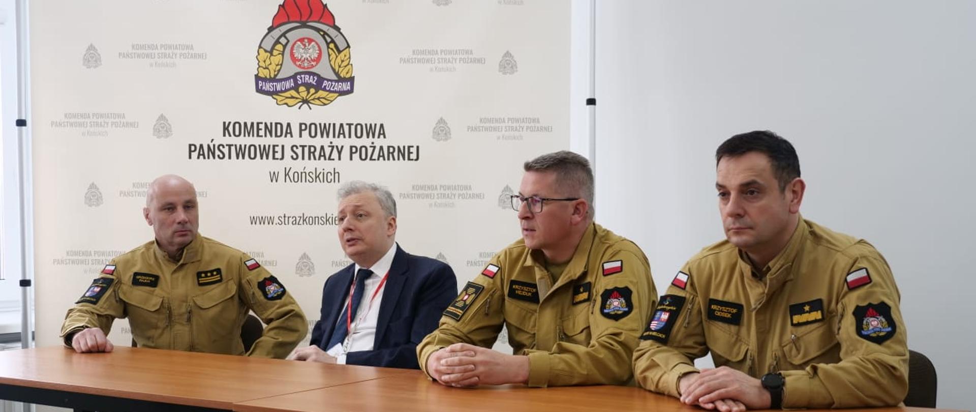 Na zdjęciu widzimy czterech mężczyzn siedzących za stołem prezydialnym w budynku. Za ich plecami ścianka z napisem KP PSP Końskie i logo PSP. Drugi od prawej jest w garniturze pozostali w mundurach piaskowych strażackich.