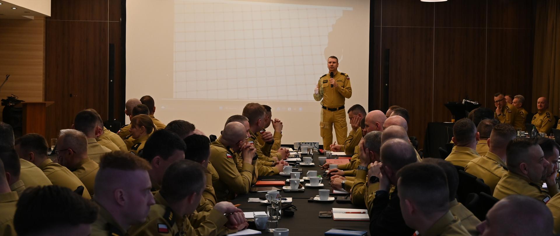 Prelegent w mundurze Państwowej Straży Pożarnej stoi przed ekranem projekcyjnym i przemawia do zgromadzonych strażaków. Uczestnicy siedzą przy stołach, skierowani w jego stronę, słuchają i notują. Na stołach widoczne są filiżanki, dokumenty oraz urządzenia elektroniczne.