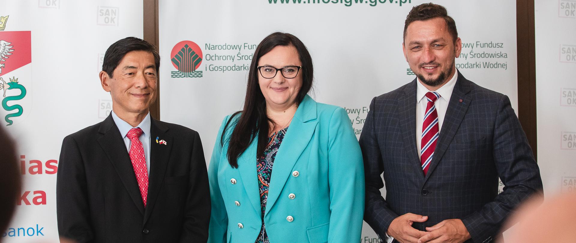 Trzy osoby pozują w sali do zdjęcia. W środku wiceminister Małgorzata Jarosińska-Jedynak. Obok burmistrz Sanoka i Ambasador Japonii.