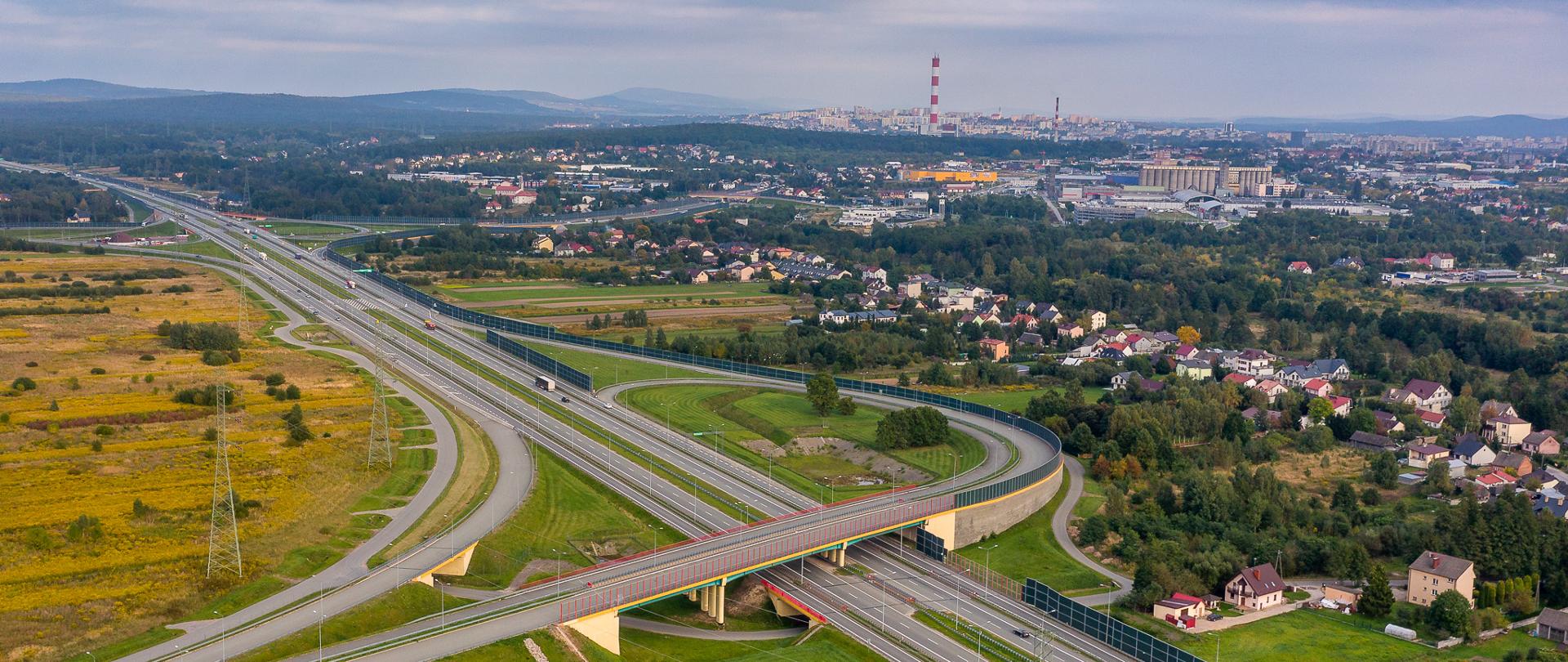 Węzeł Kielce Zachód - zdjęcie z drona, węzeł zespolony na dwujezdniowej obwodnicy Kielc w ciągu drogi ekspresowej S7. Na pierwszym planie łącznica do przyszłej drogi ekspresowej S74 w kierunku Łodzi. Dalej łącznice na skrzyżowaniu S7 z DK74, w przyszłości będzie to początek przejścia przez Kielce S74. W oddali panorama Kielc. 