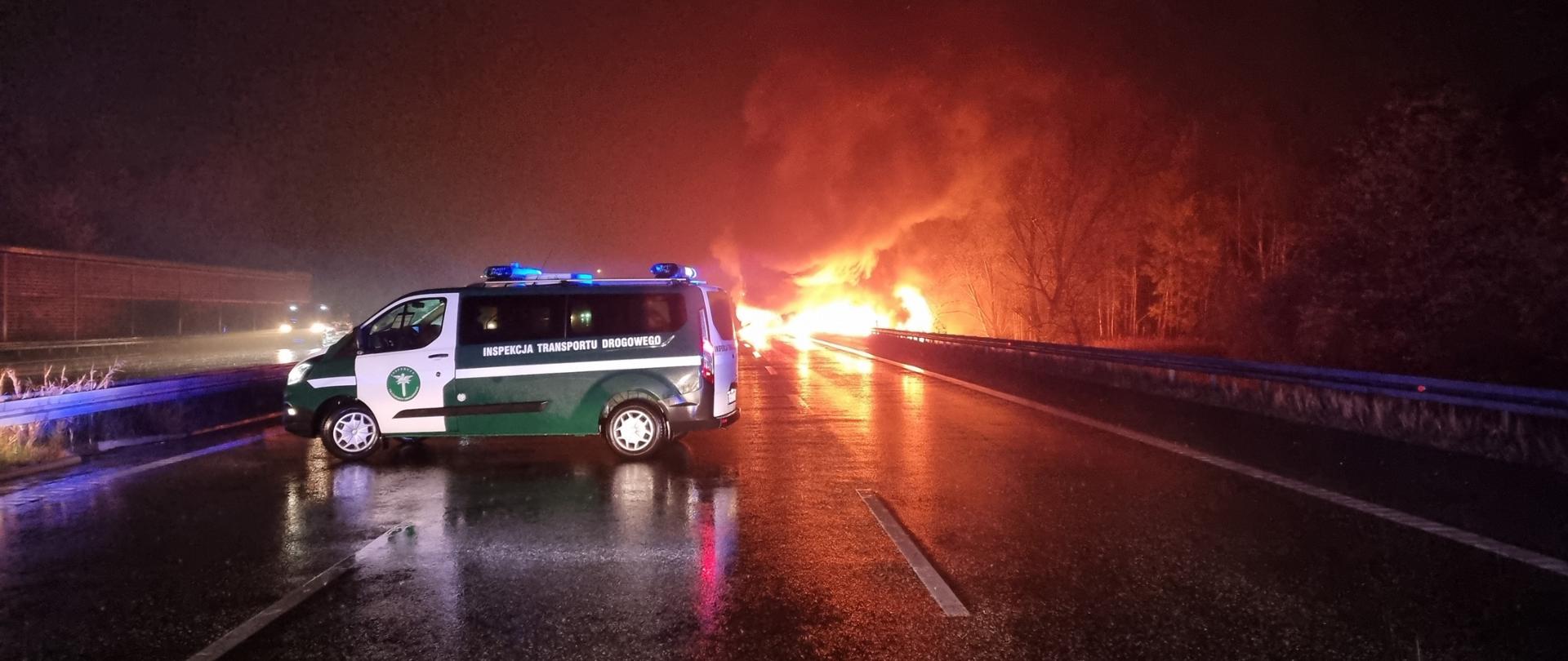 Na pierwszym planie: oznakowany radiowóz Inspekcji Transportu Drogowego stoi w poprzek jezdni i zabezpiecza miejsce śmiertelnego wypadku na autostradzie A4 w Zabrzu. W tle: płomienie i dym unoszący się nad palącą się ciężarówką.