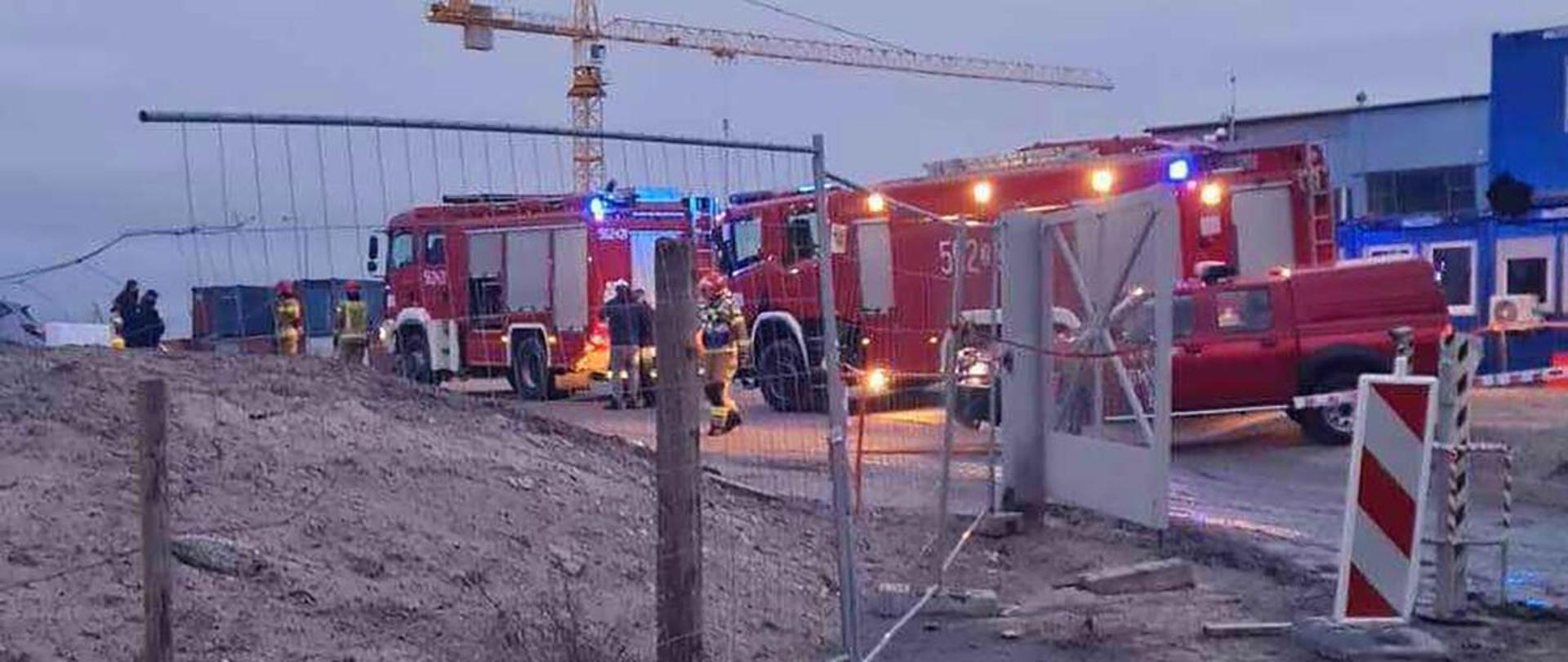 Zdjęcie przedstawia wozy straży pożarnej na placu budowy.W tle widać dźwig. Z przodu plac ogrodzony plac ogrodzony jest płotem tymczasowym. przy pojazdach widać strażaków.