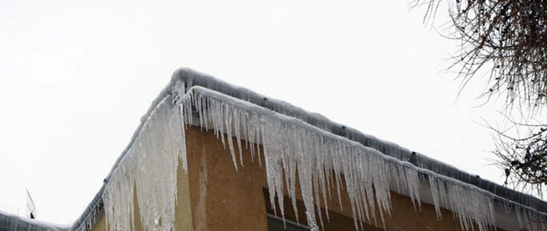 Zdjęcie przedstawia sople lodu zwisające z krawędzi dachu budynku i rynien