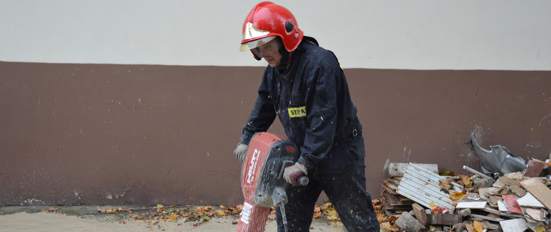 Strażak przy pomocy młota pneumatycznego rozbija beton, w celu wykonania kanału, którym przebiegać będzie instalacja wodna. Na głowie ma czerwony hełm z opuszczonymi okularami. W tle ściana budynku, po prawej stronie leżące odpady.