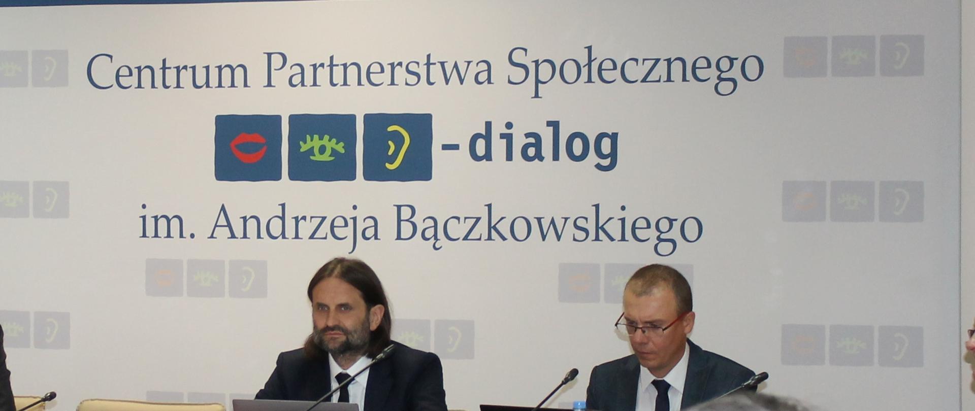 Zdjęcie przedstawia stół prezydialny za którym siedzą: Piotr Bromber - Podsekretarz Stanu w Ministerstwie Zdrowia, Współprzewodniczący Zespołu ze strony rządowej, Pan Jakub Bydłoń – Dyrektor Departamentu Dialogu Społecznego w Ministerstwie Zdrowia. W tle siedzących znajduje się baner z napisem Centrum Partnerstwa Społecznego Dialog im. Andrzeja Bączkowskiego. Nad banerem widnieje Godło Polski. 
