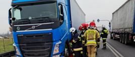 Widać samochód ciężarowy z naczepą znajdujący się na pasie awaryjnym drogi, kilku pracujących strażaków, z prawej strony przejeżdża samochód ciężarowy