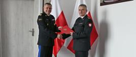 Pomorski komendant wojewódzki Państwowej Straży Pożarnej ściska dłoń oraz wręcza czerwoną teczkę. Funkcjonariusze Państwowej Straży Pożarnej ubrani w mundury wyjściowe z białą koszulą. Za nimi ustawione są dwie flagi Polski na ścianie zawieszony jest obraz.