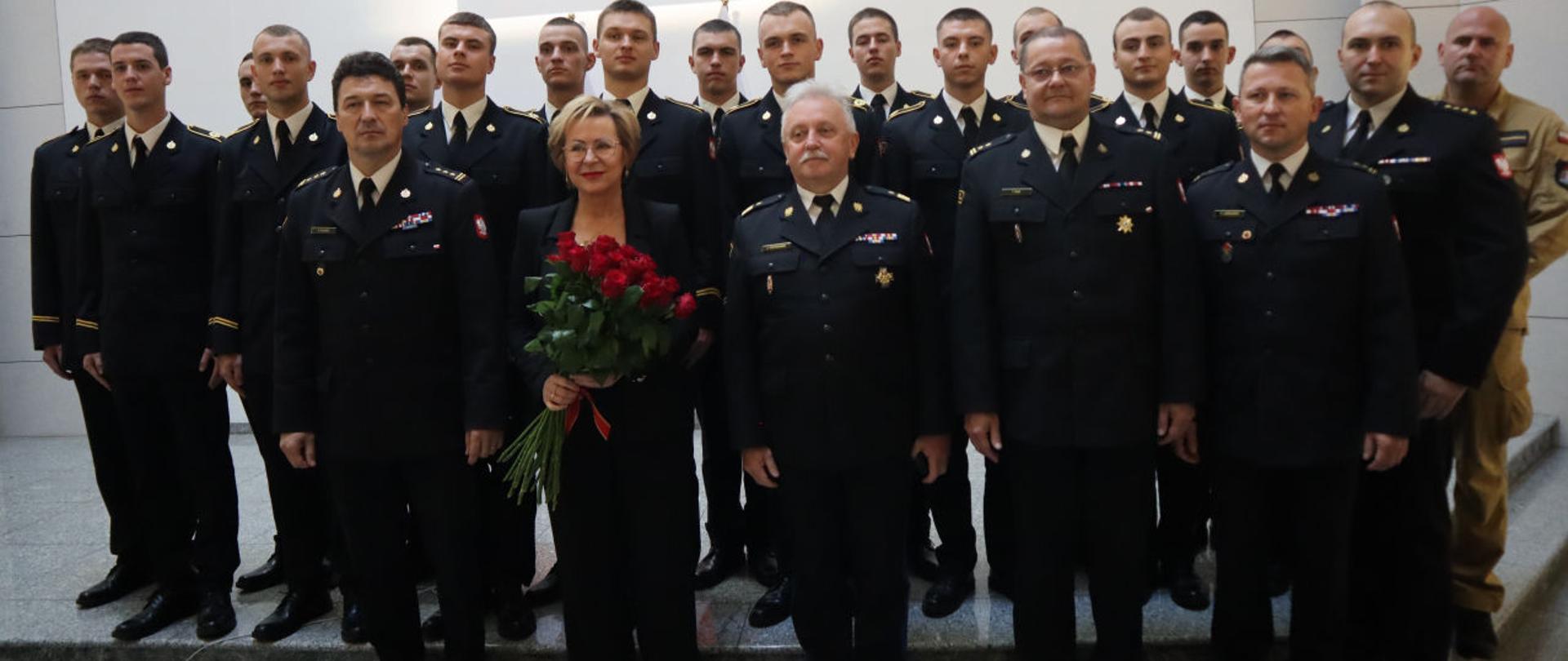 Europosłanka Jadwiga Wiśniewska na pamiątkowym zdjęciu w towarzystwie Komendanta Centralnej Szkoły PSP, Mazowieckiego Komendanta Wojewódzkiego PSP, Zastępcy Komendanta Centralnej Szkoły PSP, dwóch oficerów PSP oraz stojących z tyłu grupy kadetów CS PSP