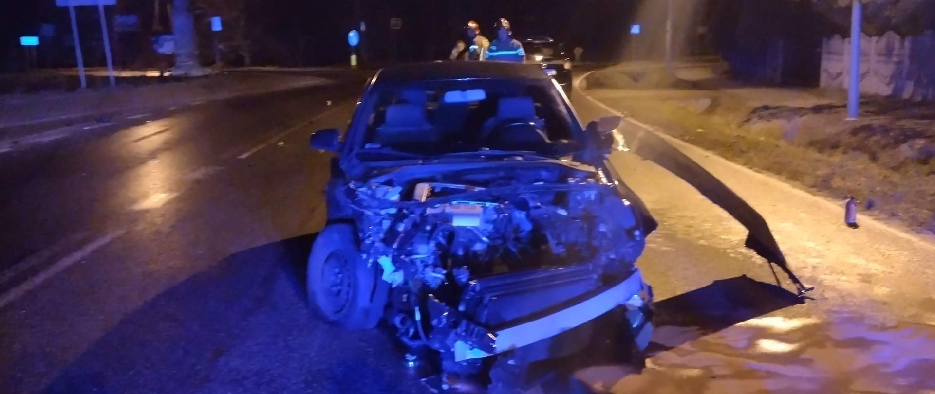 Uszkodzony samochód osobowy - zniszczony przód pojazdu. Za pojazdem 2 strażaków zabezpieczających teren działań. 