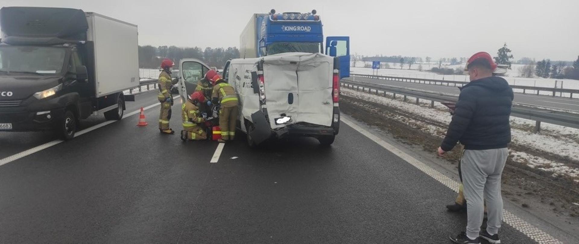 Na drodze ekspresowej stoją samochód ciężarowy i dostawczy po zderzeniu. Strażacy udzielają pomocy medycznej kierowcy furgonetki. 