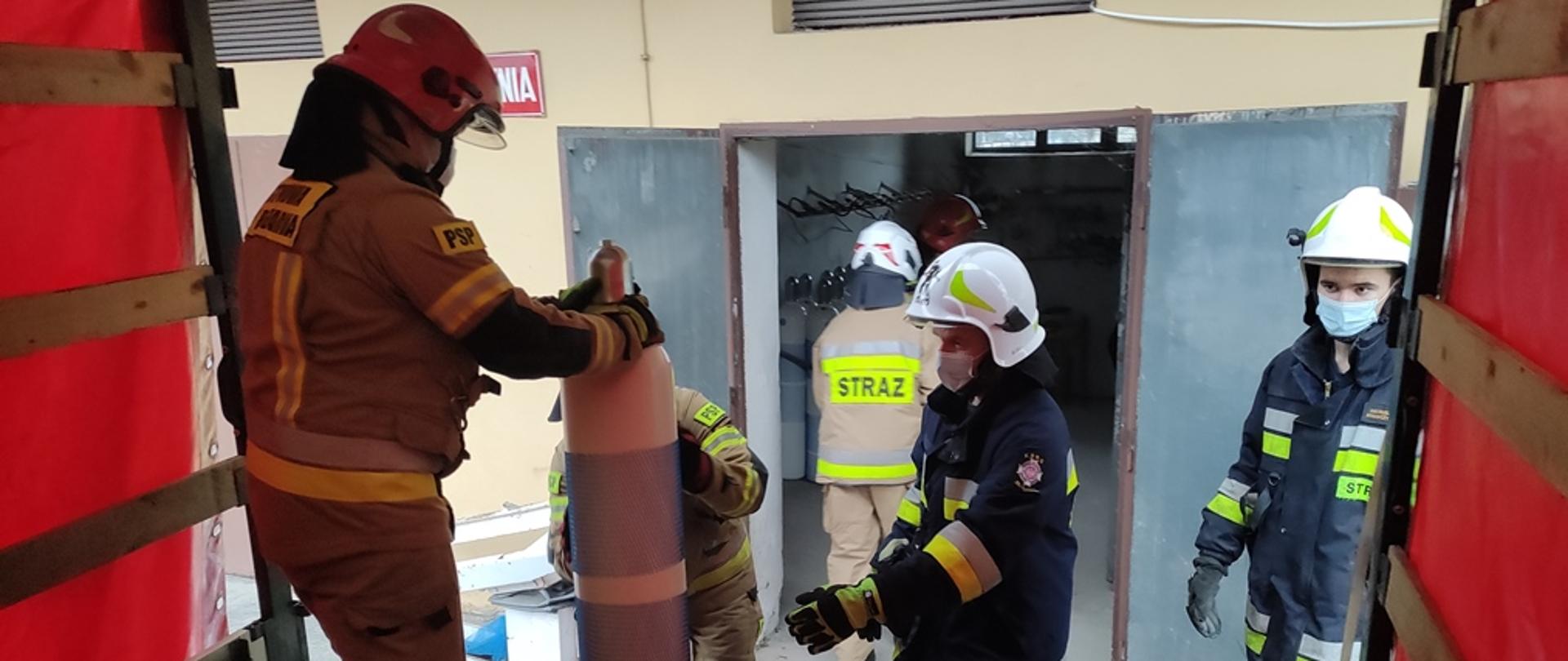 Na pierwszym planie strażaka przemieszczający butle z tlenem na pojeździe pożarniczym, na drugim palnie tlenownia i rampa rozładunkowa
