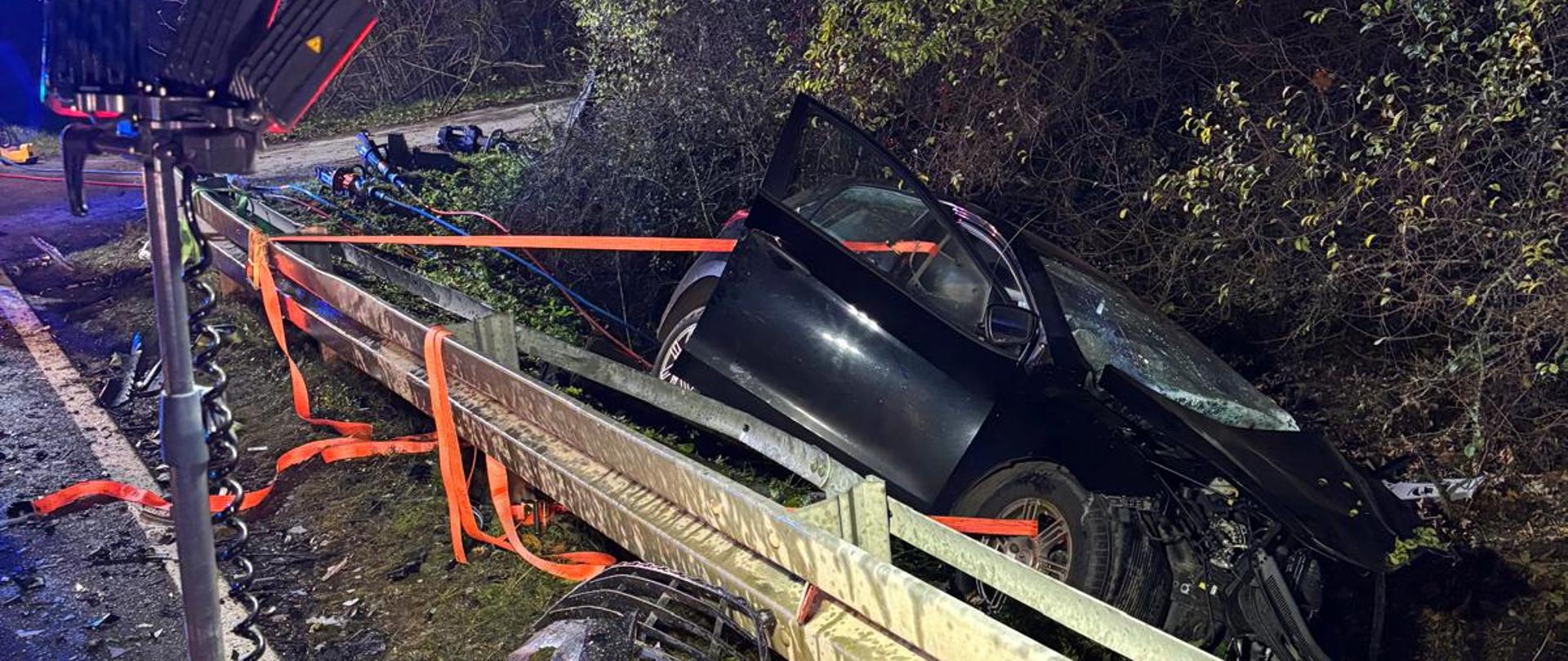 Samochód osobowy leży w rowie na boku, zabezpieczony pasami do bariery drogowej