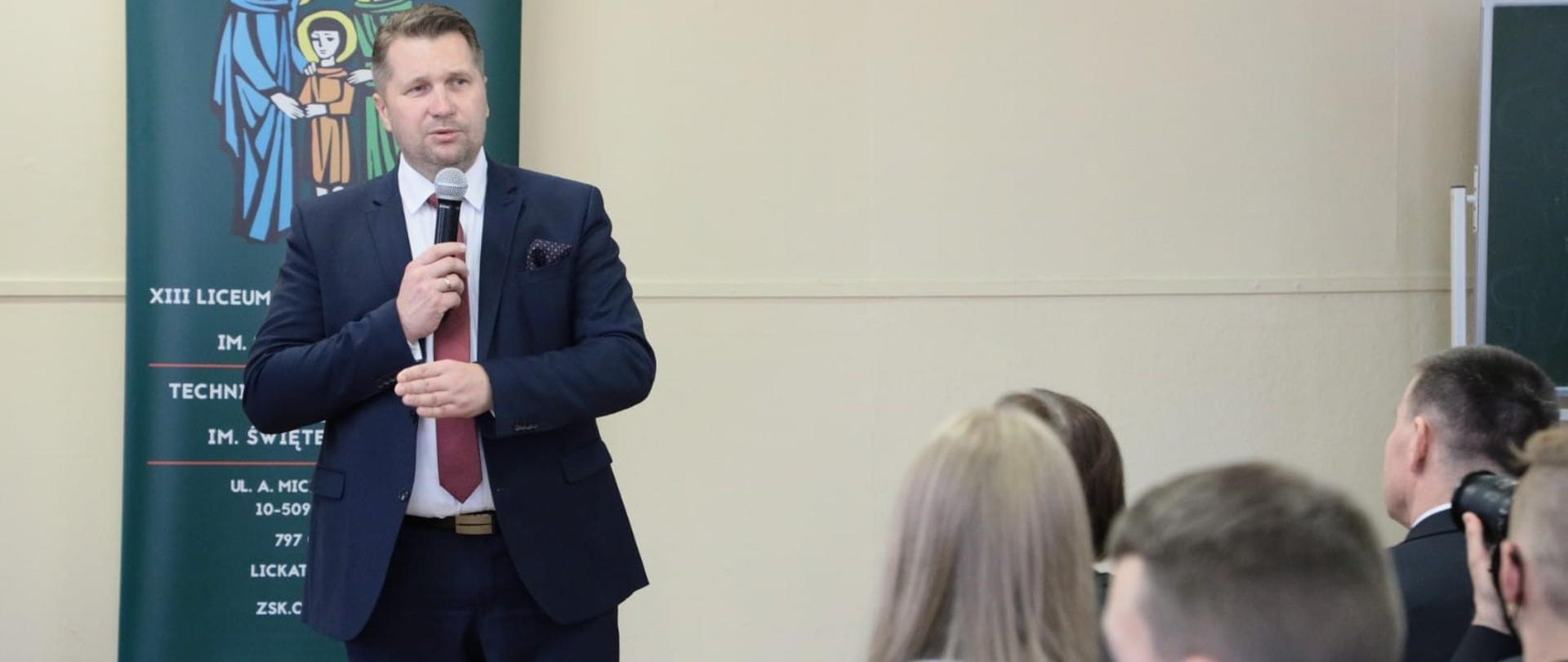Minister Przemysław Czarnek przemawia do uczniów podczas spotkania. Minister w prawej ręce trzyma mikrofon, z tyłu widoczny jest fragment roll up szkoły.
