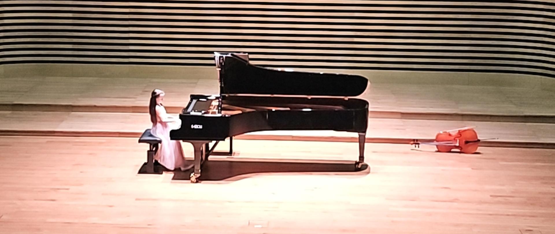 Dziewczynka w długiej jasnoróżowej sukience gra na fortepianie na estradzie sali koncertowej. Z przodu widać od tyłu fragment widowni z publicznością.