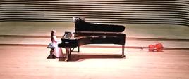 Dziewczynka w długiej jasnoróżowej sukience gra na fortepianie na estradzie sali koncertowej. 