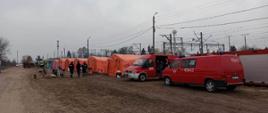 Strażacy OSP pomagają uchodźcom z Ukrainy. Fotografia wykonana w jasny dzień, widoczne dwa czerwone samochody straży pożarnej i w tle kilku strażaków stojących przy czerwonych namiotach.