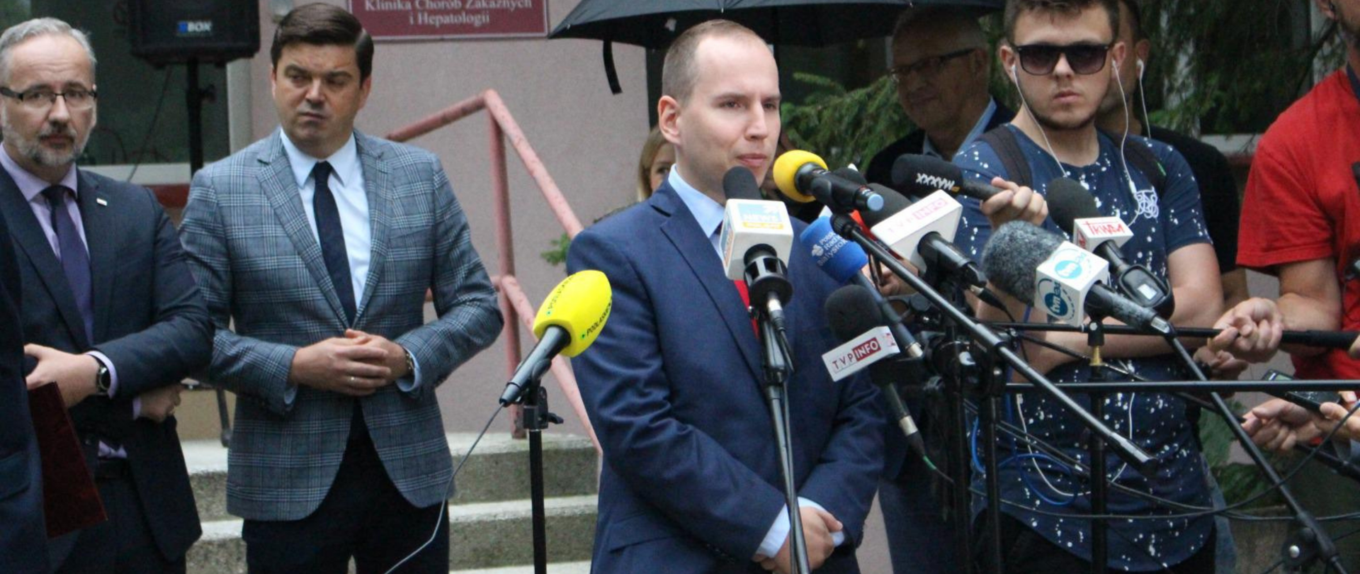 Minister Adam Andruszkiewicz w trakcie konferencji prasowej. Przed nim mikrofony dziennikarzy, z tyłu widoczny m.in. minister zdrowia Adam Niedzielski.