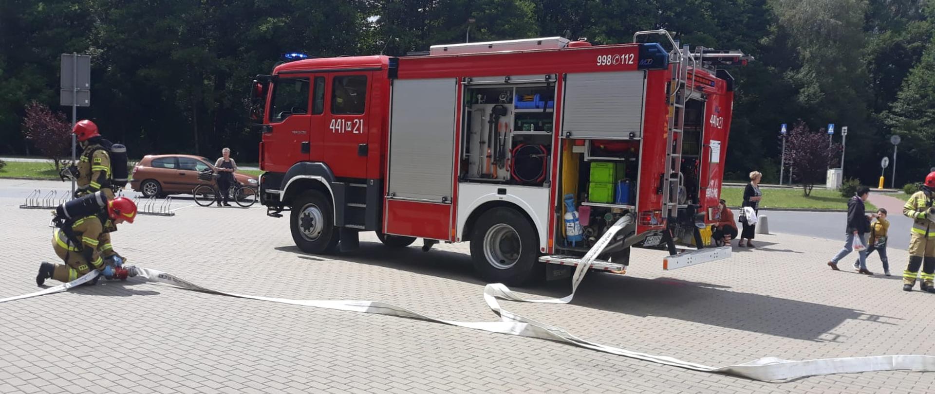 Strażacy rozwijają linię gaśniczą z węży pożarniczych która podłączona jest do samochodu pożarniczego. 