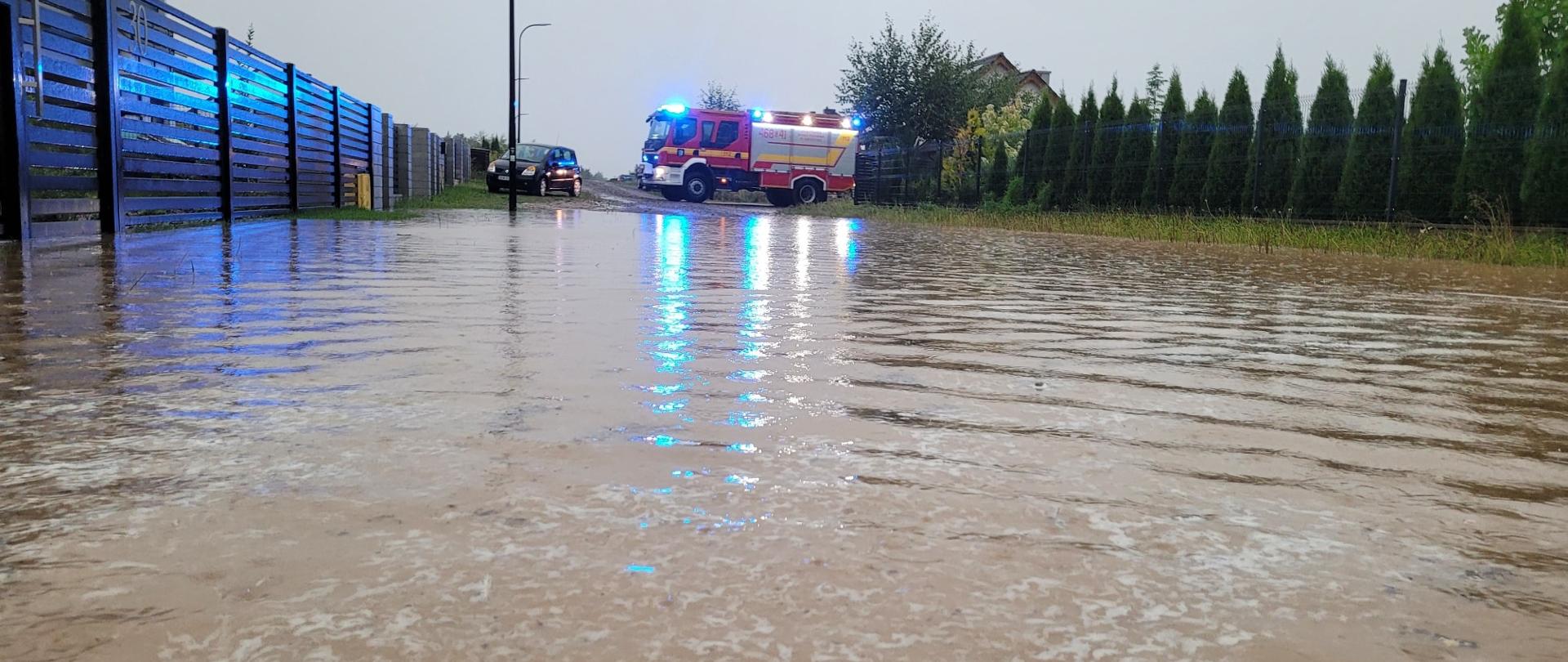 Na zdjęciu woda stojąca na drodze, w oddali samochód strażacki