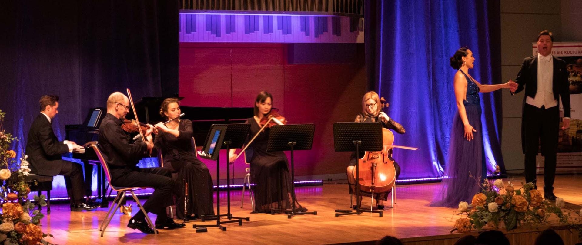 Po prawej stronie sceny ubrana w granatową suknię balową śpiewaczka, po jej lewej stronie śpiewający, ubrany we frak artysta, przed nimi scenografia składająca się z kwiatów. W tle widoczny rollup z informacjami promocyjnymi organizatorów. Za plecami kobiety widoczna niebieska poświata powstająca z podświetlenia scenicznego. Po lewej stronie orkiestra kameralna składająca się ze skrzypiec, altówki, fletu, wiolonczeli oraz fortepianu