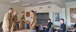Strażacy ubrani w mundury piaskowe stoją trzymając w rękach dokumenty. Dwóch strażaków siedzi przy stole ubrani są w czarne ubrania koszarowe. Wszyscy strażacy znajdują się na sali.