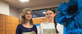 Dwie młode kobiety, Weronika Brzana i Kaja Pawlik, stoją obok siebie i trzymają w dłoniach dyplomy laureatek konkursu filmowego IPN; w tle widoczne są drewniane szafki szkolne oraz dekoracja z dużych, niebieskich kwiatów.