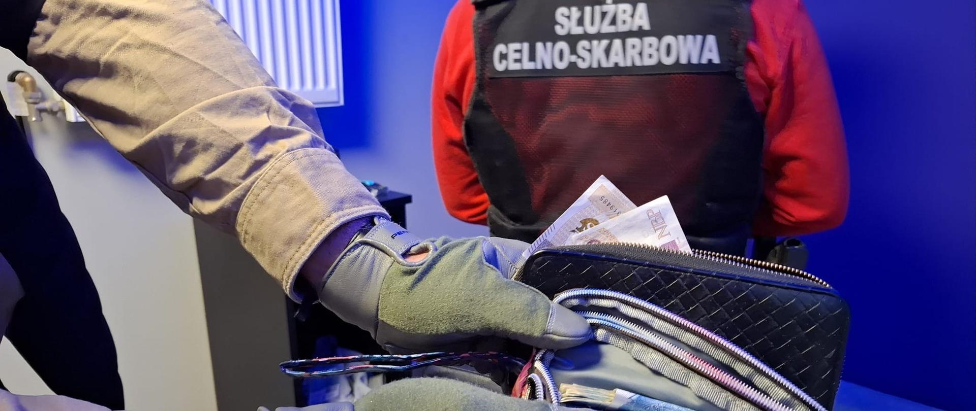 Funkcjonariusz Służby Celno-Skarbowej stoi tyłem na tle niebieskiej ściany.
Drugi otwiera saszetki z pieniędzmi.