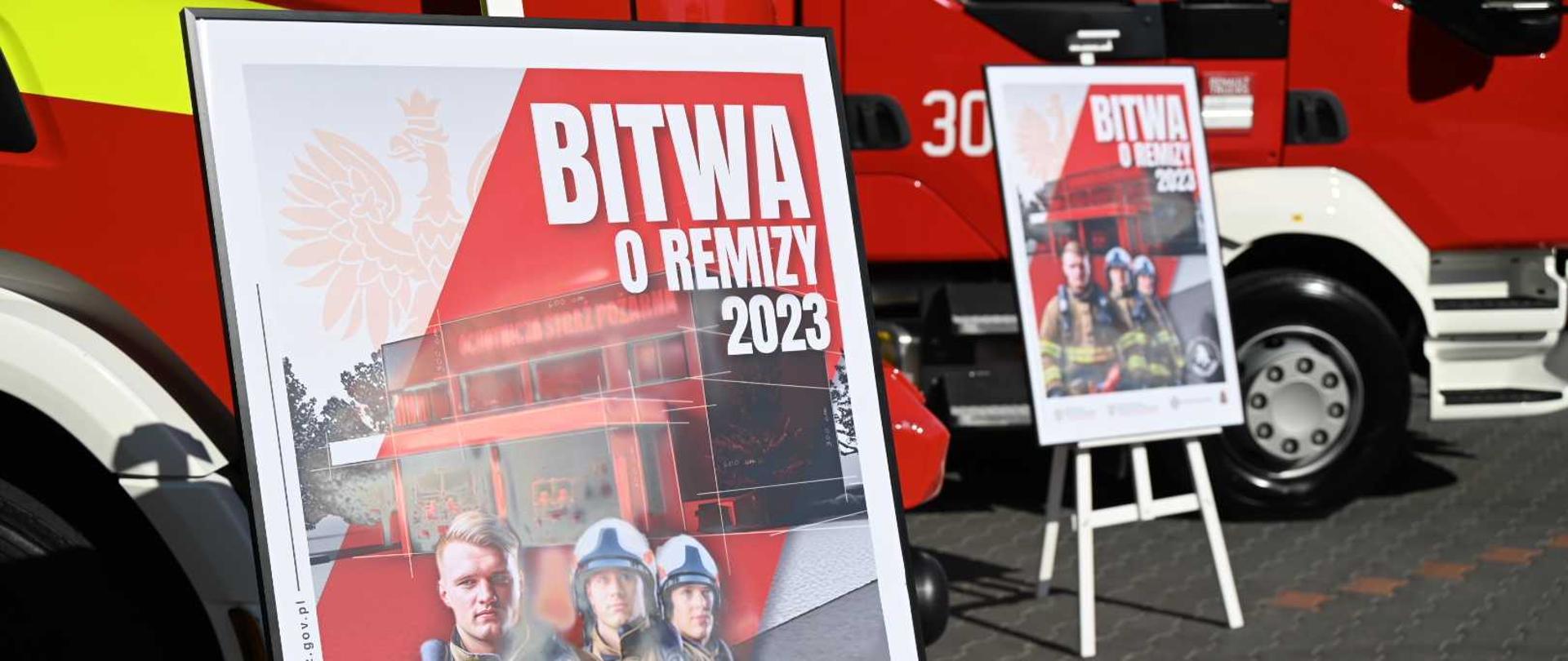 Plakaty programu „Bitwa o remizy” na tle pojazdów pożarniczych.