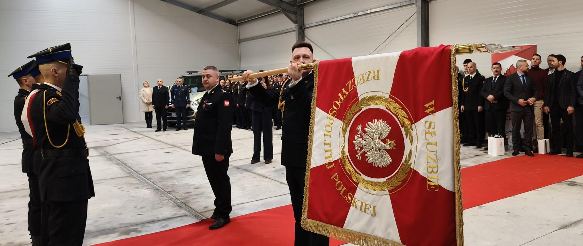 Na środku hali, gdzie odbywa się uroczystość, stoi strażak i w reku trzyma sztandar w pozycji poziomej. Obok Niego drugi strażak. W tle kilkudziesięcioosobowa załoga i goście. Dwaj pierwszoplanowi strażacy stoją na czerwonym dywanie. 