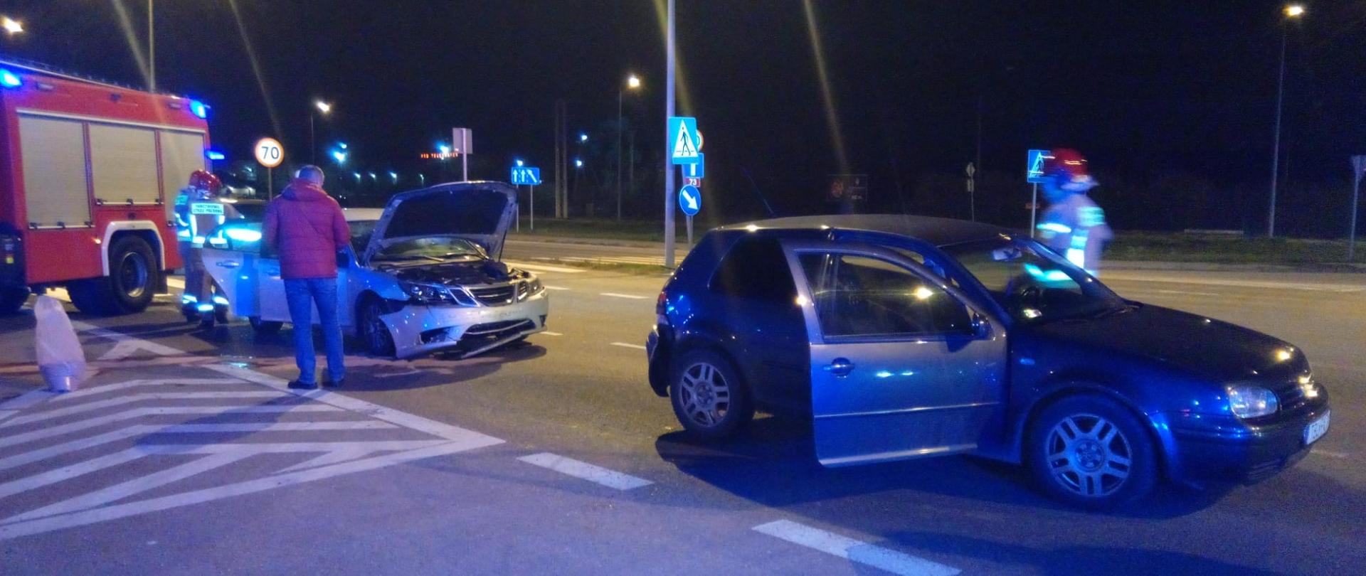 Zdjęcie przedstawia dwa samochody biorące udział w wypadku komunikacyjnym. Za nimi widać wóz strażacki. Przy drugim aucie stoi worek z sorbentem.